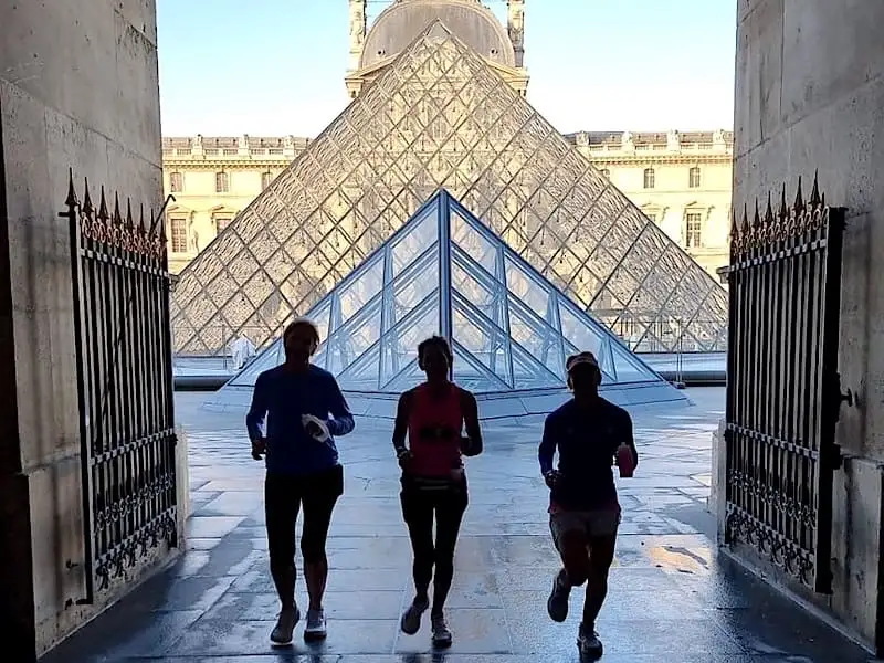 3 personas corriendo con la pirámide del Louvre de fondo